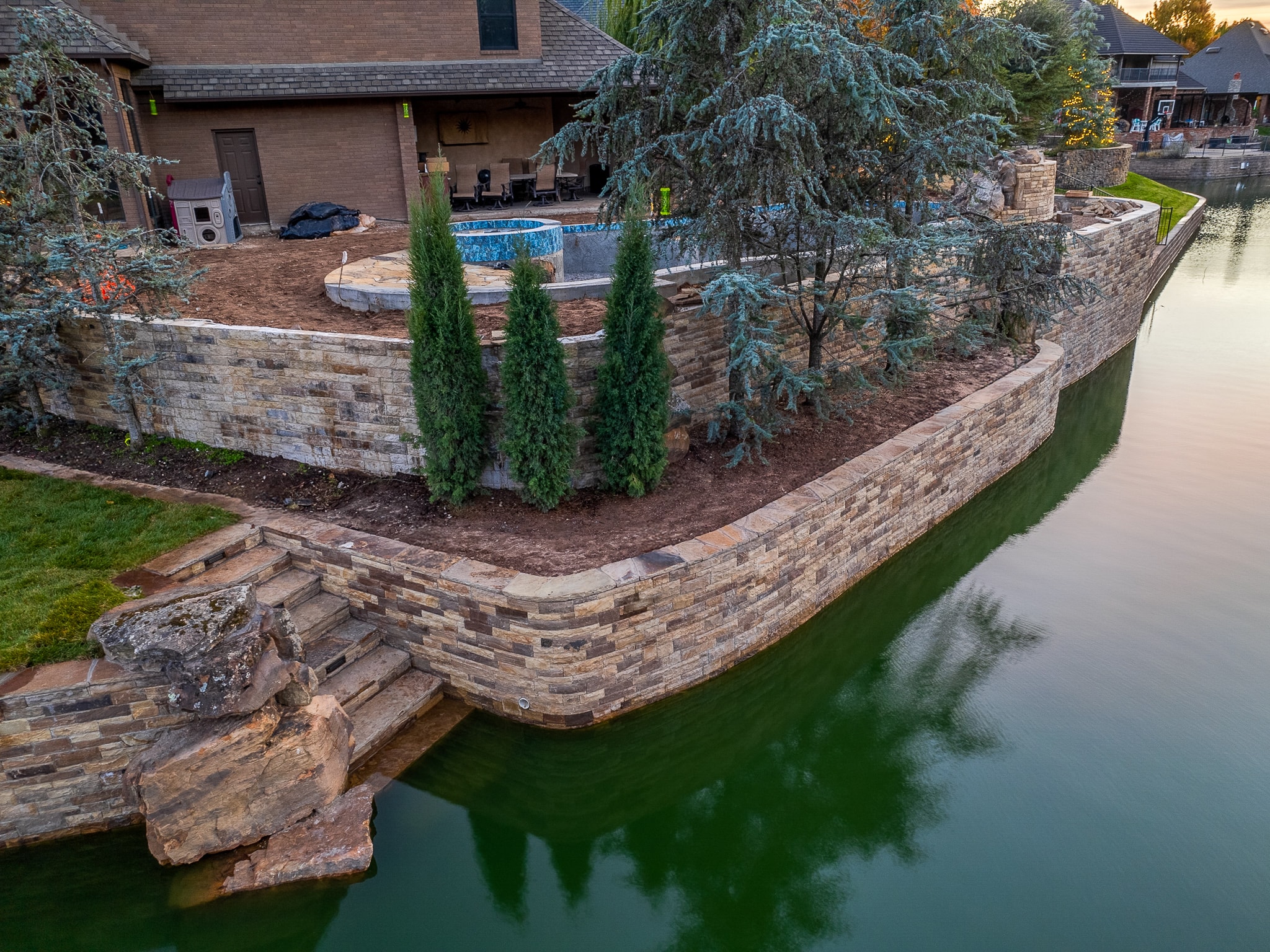 Aerial view of Stonebridge Drive lakefront stone retaining wall with Italian cypress trees and pool at dusk
