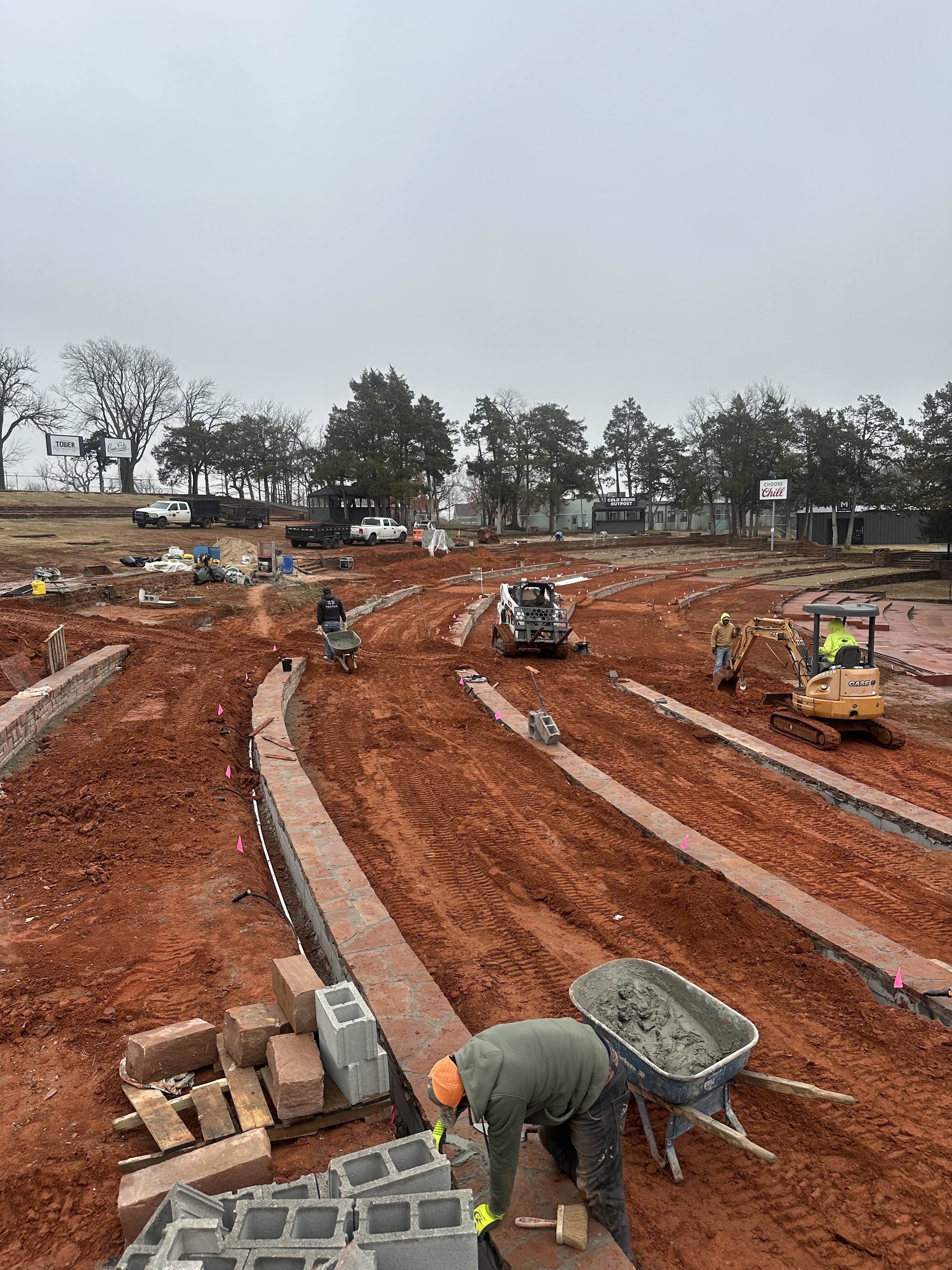 OKC Zoo Amphitheatre retaining wall construction with crew, skid steer, and excavator on red clay site