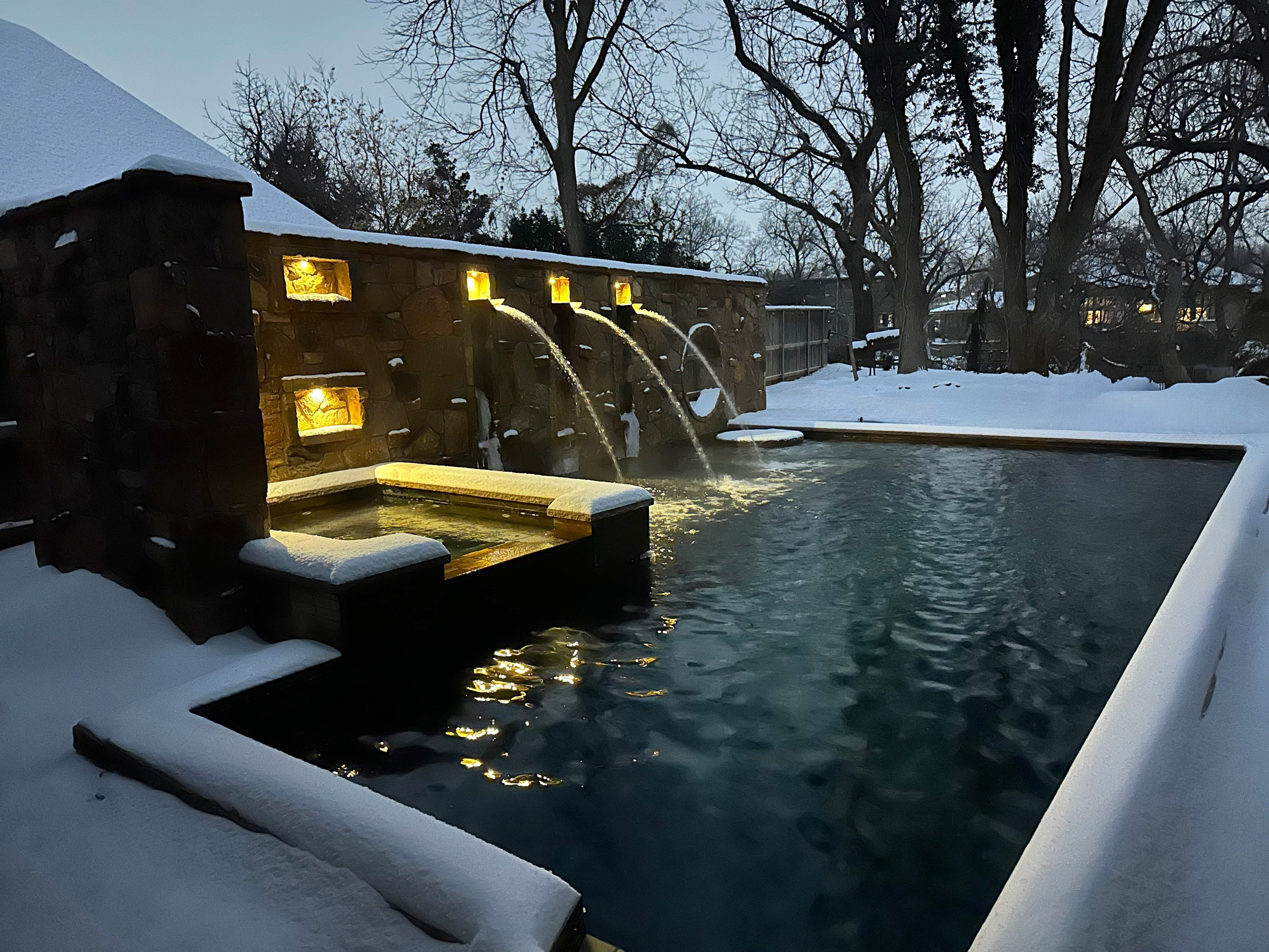 Snow-covered pool and spa with warm amber lighting and arching water jets in winter dusk